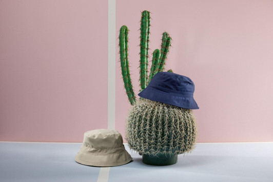 A beige bucket hat and a navy bucket hat placed on a decorative cactus against a pink background.